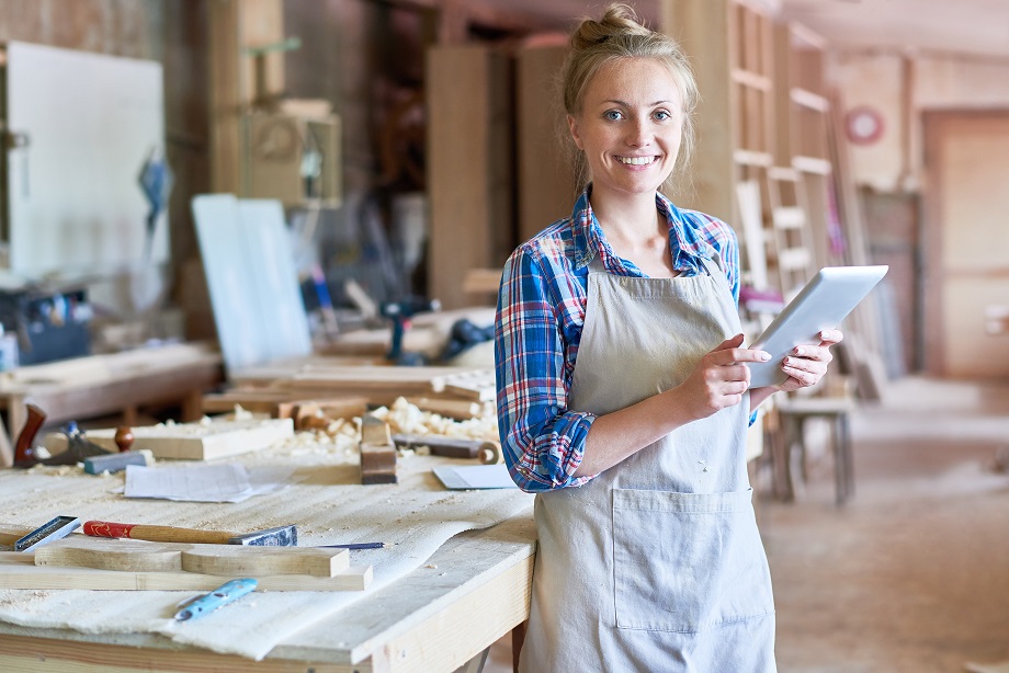 Portrait of happy young woman looking at camera using digital tablet in modern woodworking shop