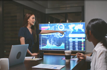 Eine junge Dame zeigt auf Daten auf einem großen Bildschirm. Ihrer Kollegin macht sich Notizen. Smart