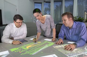 Frau mit Eyetracking-Brille und digitalem Stift zeigt ihren beiden Kollegen, über eine Projektion auf einem Tisch, ein 3D-Modell eines komplexen Bauwerkes. Digitaler Zwilling