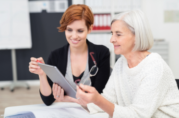 Eine junge Frau zeigt auf ein Tablet, dass ihre ältere Kollegin mit abgenommener Brille inspiziert. Innerbetriebliche Zusammenarbeit