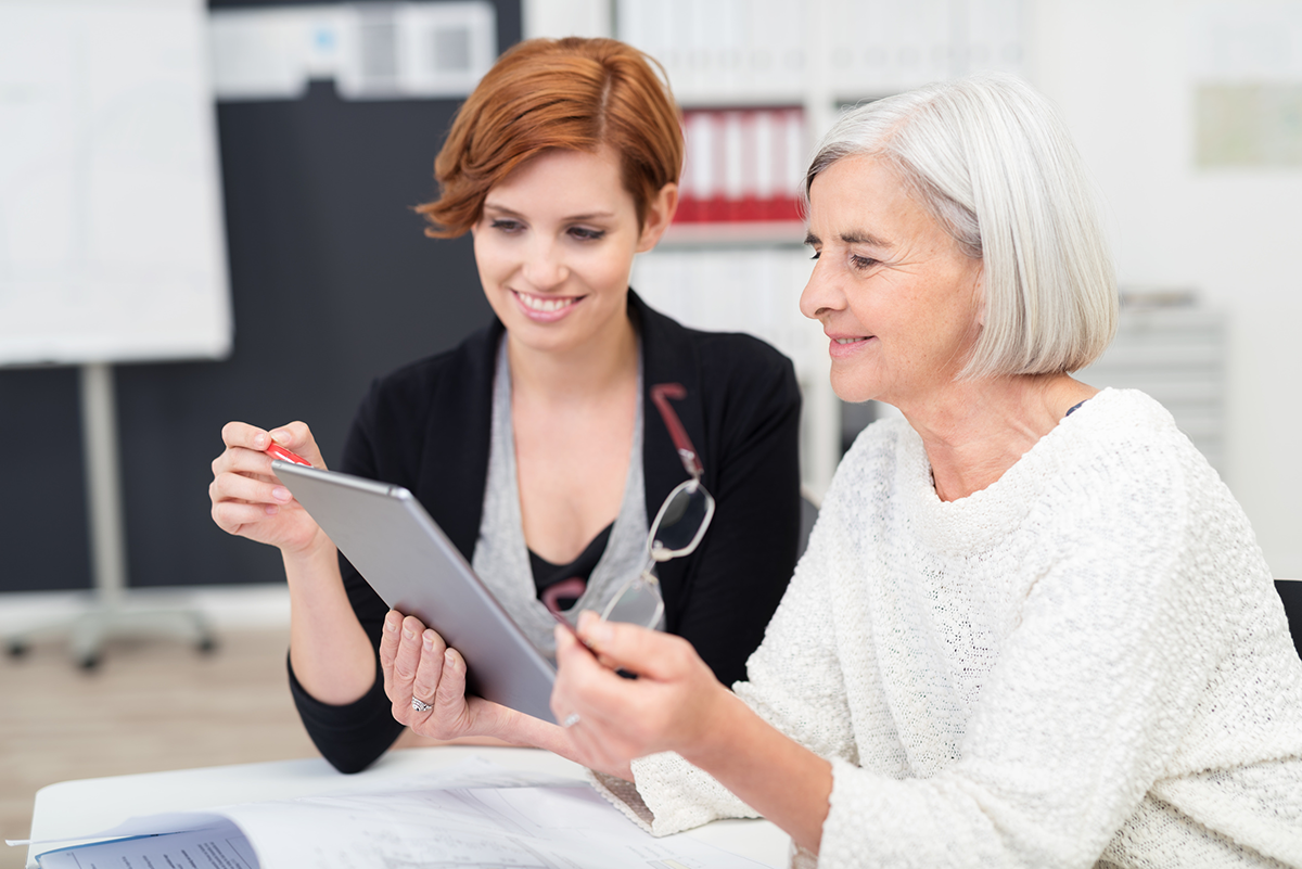 Eine junge Frau zeigt auf ein Tablet, dass ihre ältere Kollegin mit abgenommener Brille inspiziert. Innerbetriebliche Zusammenarbeit