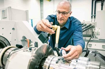 Ein älterer Mann mit Brille führt eine Abstandsmessung an einer großen Maschine durch. Handwerksarbeit