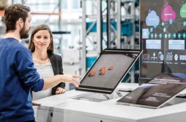 Ein Mann erklärt einer interessierten Frau den Demonstrator zu Cloud Computing in der Industrie über 3 Bildschirme