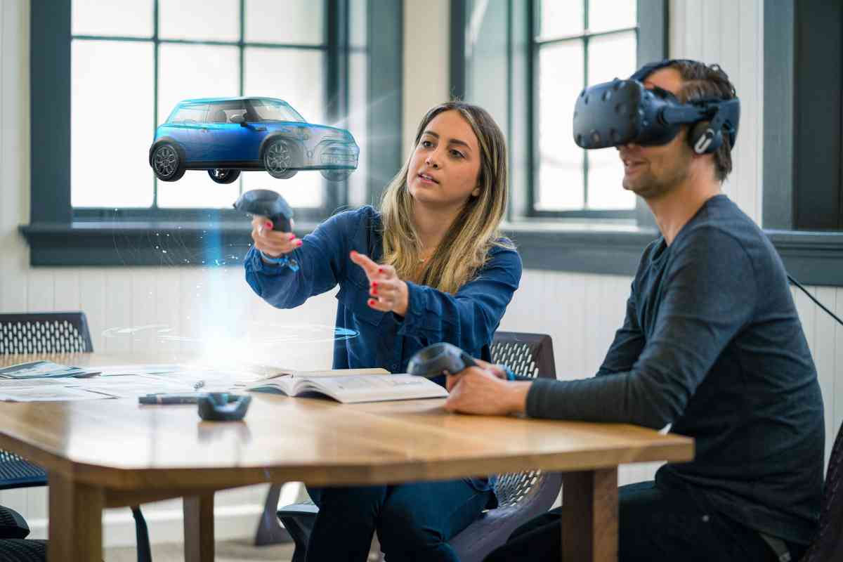 Besprechungssituation mit einer Frau und einem Mann. Er trägt eine Augmented Reality-Brille und sieht das Hologramm eines Autos.