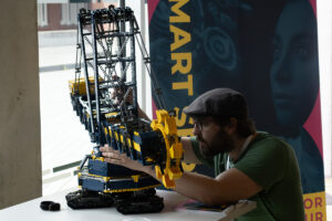 Ein Lego Modell eines Schaufelbaggers wird von einem Mann mit Baskenmütze, Bart und Brille präpariert. Smart