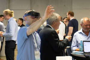 In einer Gruppe von Menschen wird eine AR-Brille benutzt. Der Mann bedient mit 2 Fingern über das virtuelle Auge