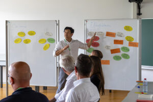 Mann der energisch auf eine Stellwand zeigt auf der beschriftete, farbige, runde und eckige Papiere kleben. Erfolgsfaktoren