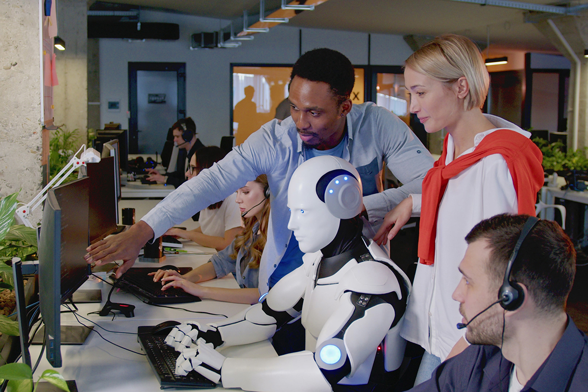 Zwei Männer und eine Frau in Besprechung in einem Call Center. Sie erklären einem humanoiden Roboter die Arbeitsschritte. Der Roboter arbeitet im Call Center.
