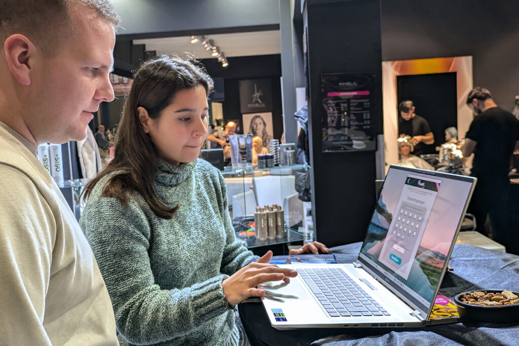Eine Frau und ein Mann stehen auf der Messe vor einem Laptop und sehen sich das Ergebnis der FrisAR App auf dem Bildschirm an.