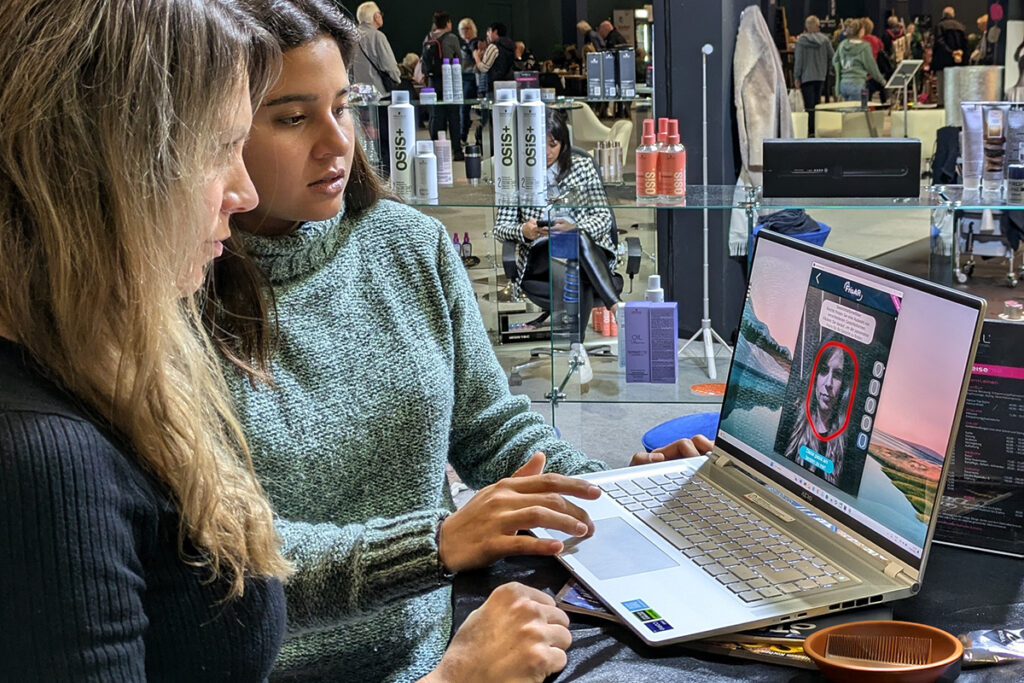 Zwei Frauen stehen auf der Messe vor einem Laptop und sehen sich das Ergebnis der FrisAR App auf dem Bildschirm an