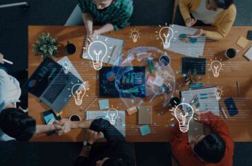 Bild der Bekanntmachung „Internationale Orte der Innovation: Herausforderungen internationaler Wertschöpfung identifizieren, Lösungen kollaborativ entwickeln, technologische Souveränität stärken“. Das Bild zeigt Frauen und Männer, die zu einer Besprechung an einem Tisch sitzen. Die Icons "Glühbirnen" und "Weltkugel" symbolisieren, dass Lösungen entwickelt werden, mit denen internationale Wertschöpfung identifiziert und technologische Souveränität gestärkt wird.