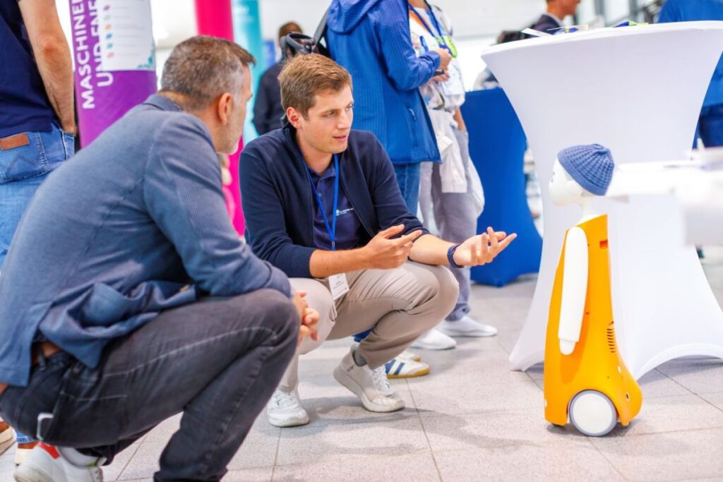 Ein Mann zeigt einem anderen Mann anhand eines Roboters ein Beispiel für die datenbasierten und KI-gestützten Assistenzsystem. Dies ist ein Forschungsergebnis des Projekts PAL.